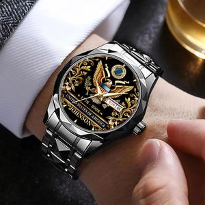 May include: A silver-toned wristwatch with a black face featuring a golden eagle design, floral accents, and the text "UNITED STATES OF AMERICA 1776-2026 JOHNSON". The watch has a silver band and is worn on a wrist.