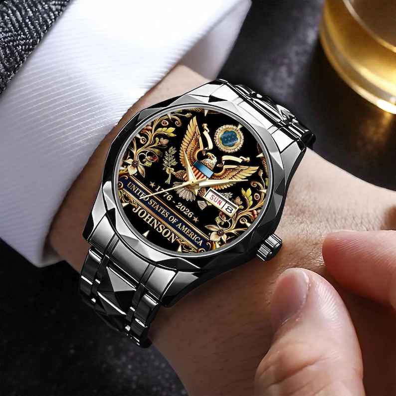 May include: A silver-toned wristwatch with a black face featuring a golden eagle design, floral accents, and the text "UNITED STATES OF AMERICA 1776-2026 JOHNSON". The watch has a silver band and is worn on a wrist.