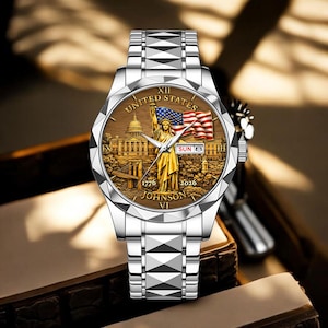 Peut inclure: Montre-bracelet argent&eacute;e avec un cadran d&eacute;taill&eacute; repr&eacute;sentant la Statue de la Libert&eacute;, un drapeau am&eacute;ricain et le texte "UNITED STATES". Le bracelet a un motif g&eacute;om&eacute;trique. La date est affich&eacute;e.