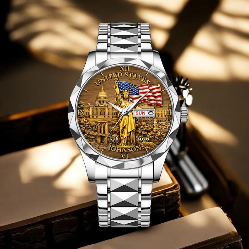 Peut inclure: Montre-bracelet argent&eacute;e avec un cadran d&eacute;taill&eacute; repr&eacute;sentant la Statue de la Libert&eacute;, un drapeau am&eacute;ricain et le texte "UNITED STATES". Le bracelet a un motif g&eacute;om&eacute;trique. La date est affich&eacute;e.