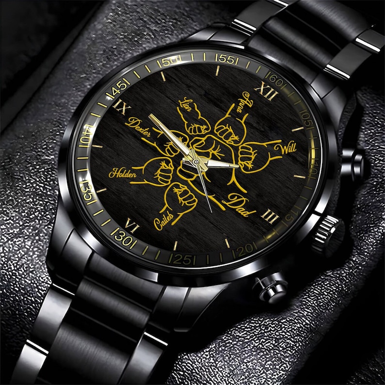 May include: Black wristwatch with a black band and a black face. The face has a wood grain pattern and gold accents. The numbers on the face are Roman numerals. The words "Dad", "Lion", "Dexter", "Anna", "Holden", "Caileb", and "Will" are written on the face in gold lettering.