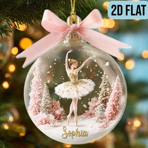 May include: A clear glass Christmas bauble with a ballerina in a gold and white tutu, surrounded by pink trees and falling snow. The bauble is adorned with a pink bow and the name "Sophia".