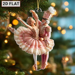 May include: A 2D flat Christmas ornament depicting a ballerina and a nutcracker in a dance pose. The ballerina wears a pink tutu and the nutcracker is in a pink uniform. The ornament is suspended from a gold string.