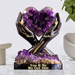 May include: A decorative sculpture featuring a heart-shaped amethyst crystal held by two black hands with gold fingernails. The base has the text "You & Me We Got This" and the names "David" and "Sophia".