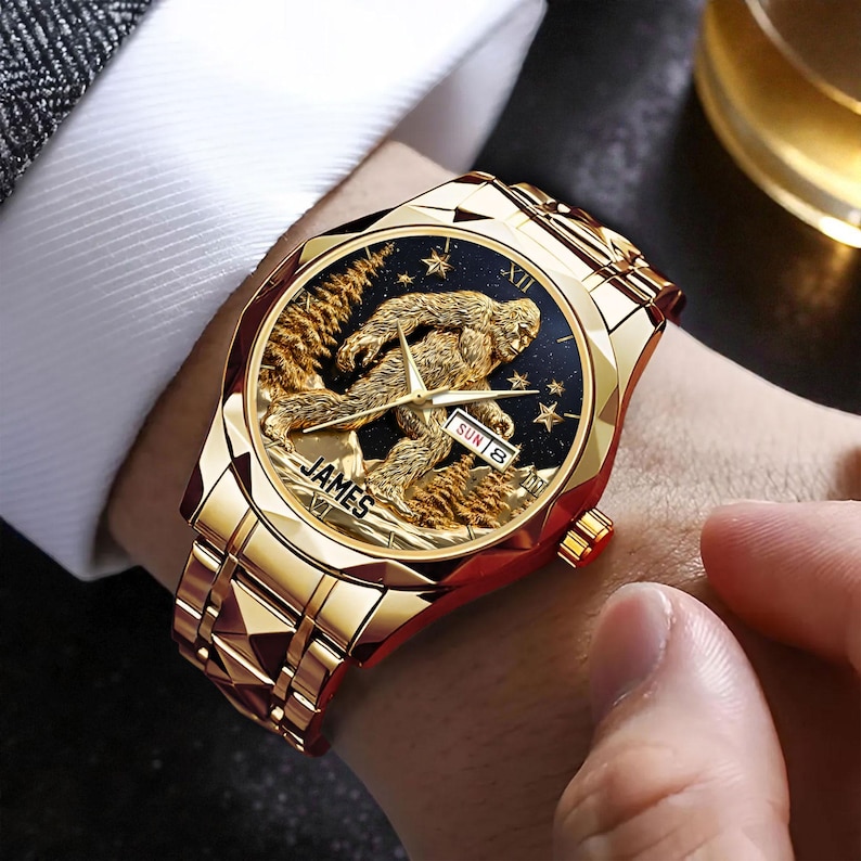 May include: A gold-toned wristwatch with a detailed Sasquatch design on the black watch face. The watch features a gold band and the word "JAMES" is visible. The day and date are displayed on the watch face.