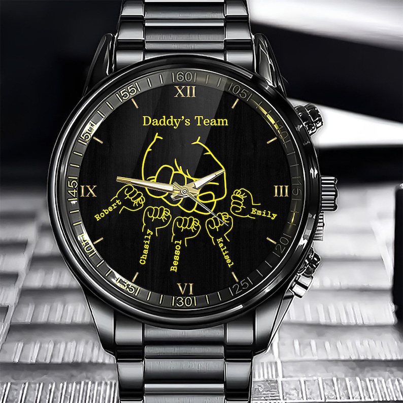 May include: Black wristwatch with a black face and gold accents. The face has the words "Daddy's Team" and the names "Robert", "Chasily", "Bessol", "Kalisel", and "Emily" around the edge.