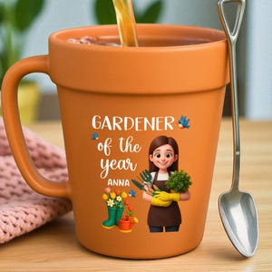 May include: Orange ceramic mug shaped like a flower pot, with the text "GARDENER of the year ANNA" and a cartoon illustration of a gardener. A silver spoon rests beside the mug. The mug is filled with a liquid.