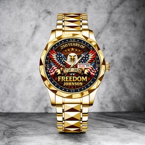 Puede incluir: Un reloj dorado con un dise&ntilde;o patri&oacute;tico. La esfera del reloj presenta un &aacute;guila, banderas americanas y el texto "250 Years of Freedom Johnson 1776-2026". La correa del reloj tiene un patr&oacute;n geom&eacute;trico.