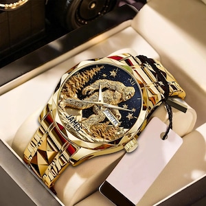 May include: Gold-toned wristwatch with a detailed Sasquatch design on the face. The watch features a gold band and a black background with stars. The watch is in a presentation box, with a tag attached.