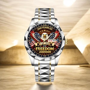 Puede incluir: Reloj de pulsera plateado y dorado con un dise&ntilde;o patri&oacute;tico. La esfera del reloj presenta un &aacute;guila, banderas americanas y el texto "250 YEARS OF FREEDOM JOHNSON 1776-2026". La correa del reloj es plateada y dorada.