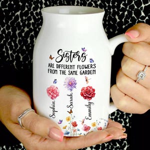 Puede incluir: Taza de cerámica blanca con asa, con el texto "Sisters" y un diseño floral con los nombres Sophia, Sarah y Emmily. La taza también tiene mariposas y la cita "ARE DIFFERENT FLOWERS FROM THE SAME GARDEN."