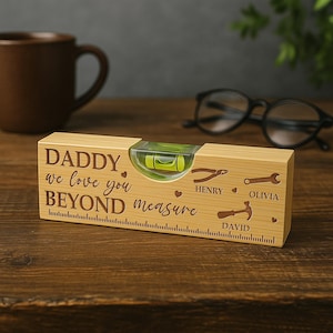 Puede incluir: Un nivel de madera con el texto "DADDY we love you BEYOND measure" y los nombres Henry, Olivia y David. El nivel tiene una burbuja y una regla en la parte inferior. Una taza marrón y gafas están en el fondo.