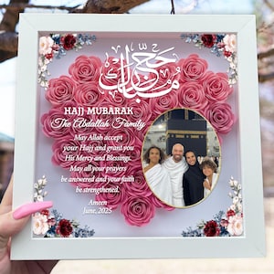 May include: A white framed artwork with a heart-shaped arrangement of pink paper roses. The artwork includes a photo of a family and text that reads "HAJJ MUBARAK" and "The Abdallah Family". Decorative floral accents are in the corners.