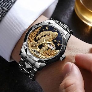 May include: A silver-toned wristwatch with a black face featuring a gold-colored Sasquatch design. The watch face also displays the day and date. The watch has a silver band and the word "JAMES" is printed on the watch face.