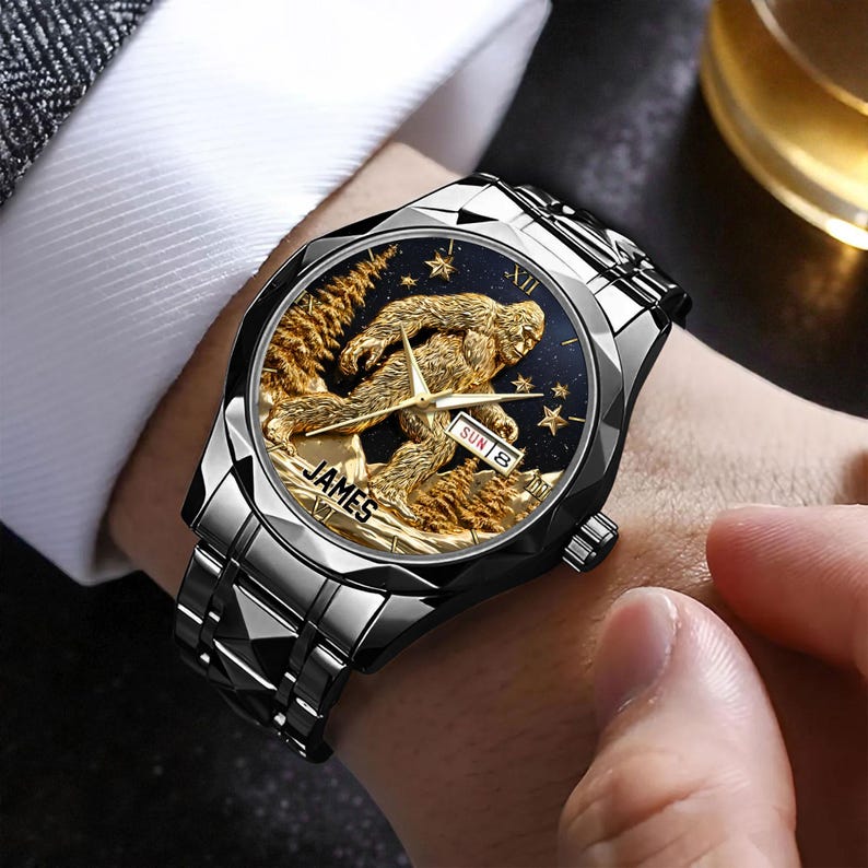 May include: A silver-toned wristwatch with a black face featuring a gold-colored Sasquatch design. The watch face also displays the day and date. The watch has a silver band and the word "JAMES" is printed on the watch face.