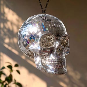 May include: A decorative skull-shaped disco ball, hanging from a black chain. The skull is covered in small, reflective mirrored tiles that catch the light, creating a dazzling effect. The skull's eye sockets and nose cavity are visible.