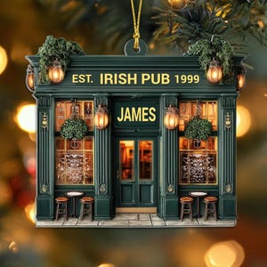 May include: A green ornament depicting an Irish pub facade, with the text "EST. IRISH PUB 1999" and "JAMES". The pub features lanterns, small tables, and stools. The ornament is suspended by a gold string.