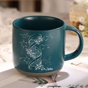 May include: A teal blue ceramic mug with a white design of a DNA strand with flowers and chemical formulas. The mug has a rounded handle and the text "Dr. Sophia" is printed on the bottom.