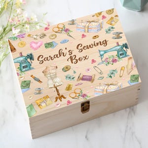 May include: A wooden box with a hinged lid and a latch. The box is decorated with a colorful pattern of sewing-themed illustrations, including a sewing machine, fabric scraps, buttons, and needles. The text "Sarah's Sewing Box" is printed on the lid.