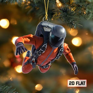 May include: A 2D flat Christmas ornament featuring a person in an orange jumpsuit and black helmet, with a parachute pack. The ornament has the text "DAVID-2025" and is hanging from a gold string. The words "2D FLAT" are at the bottom.