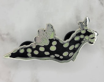 Puede incluir: Un pin de esmalte negro y plateado con forma de babosa de mar con un diseño de mariposa. El pin presenta un cuerpo negro con lunares y detalles en verde claro. Las alas y antenas de la mariposa son plateadas.