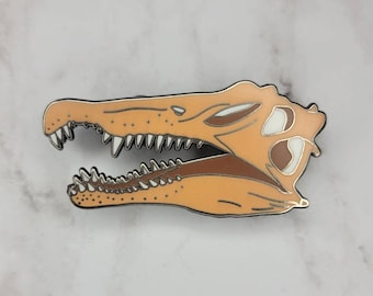 Puede incluir: Un pin de esmalte metálico con un detallado cráneo de dinosaurio naranja y blanco con dientes afilados. El pin es una gran adición a la colección de cualquier entusiasta de los dinosaurios.