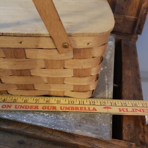 Puede incluir: Una cesta de picnic de madera con un dise&ntilde;o tejido y una tapa con bisagras. La cesta tiene un asa de madera y est&aacute; sentada sobre una superficie de madera. El texto "ON UNDER OUR UMBRELLA" est&aacute; impreso en una regla.