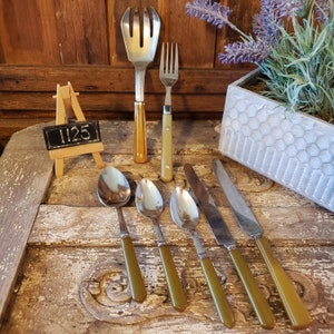 Könnte beinhalten: Ein Set aus sechs silbernen Besteckteilen, darunter eine Gabel, ein Schöpflöffel und vier Löffel, mit olivgrünen Griffen. Das Besteck ist auf einer Holzoberfläche mit einem kleinen Schild mit der Aufschrift "1125" angeordnet.