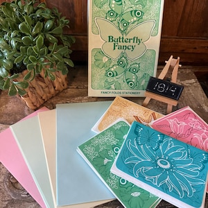 May include: A stationery set with a butterfly theme. The set includes a box of "Butterfly Fancy" stationery, along with several pastel-colored blank cards and floral-patterned cards in various colors, including pink, green, and teal. A small chalkboard displays the number 1814.