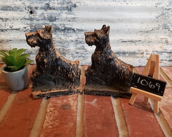 May include: A pair of black ceramic bookends shaped like Scottish Terriers. The bookends are sitting on a brick surface with a small chalkboard easel in front of them. The chalkboard has the number 1069 written on it.