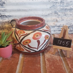 Puede incluir: Una maceta de cerámica blanca con un borde marrón y diseños florales rojos, naranjas y negros. La maceta tiene un asa y está sentada sobre una superficie de madera. Un pequeño caballete de pizarra con el número 1155 escrito en él está en el fondo.