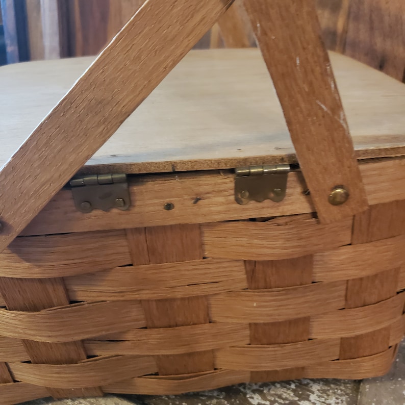 Puede incluir: Una cesta de picnic de madera con tapa con bisagras y base de cesta tejida. La cesta tiene dos bisagras de metal y un cierre de lat&oacute;n.