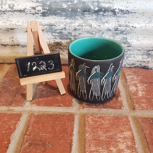 Puede incluir: Un macetero de cerámica negra con un borde verde azulado presenta un diseño gráfico blanco de figuras estilizadas bailando alrededor del macetero. El macetero está sentado sobre una superficie de ladrillo con un pequeño caballete de madera y una pizarra que dice "123".