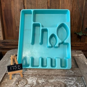 Puede incluir: Una bandeja de cubiertos de plástico turquesa con múltiples compartimentos para organizar la cubertería. La bandeja tiene ranuras para cuchillos, tenedores y cucharas, y está diseñada para encajar dentro de un cajón de cocina. Un pequeño caballete de madera con el número "1755" está en primer plano.