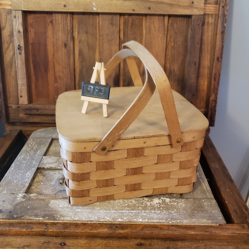 Puede incluir: Una cesta de picnic de madera vintage con tapa tejida y asa de cuero marr&oacute;n. La cesta est&aacute; sobre una superficie de madera con un peque&ntilde;o caballete de pizarra en la parte superior.