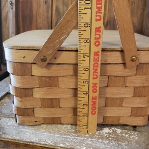 Puede incluir: Una cesta de picnic de madera con tapa abatible y dise&ntilde;o tejido. La cesta tiene dos asas y una regla a su lado que dice "Come on Under Our Umbrella".