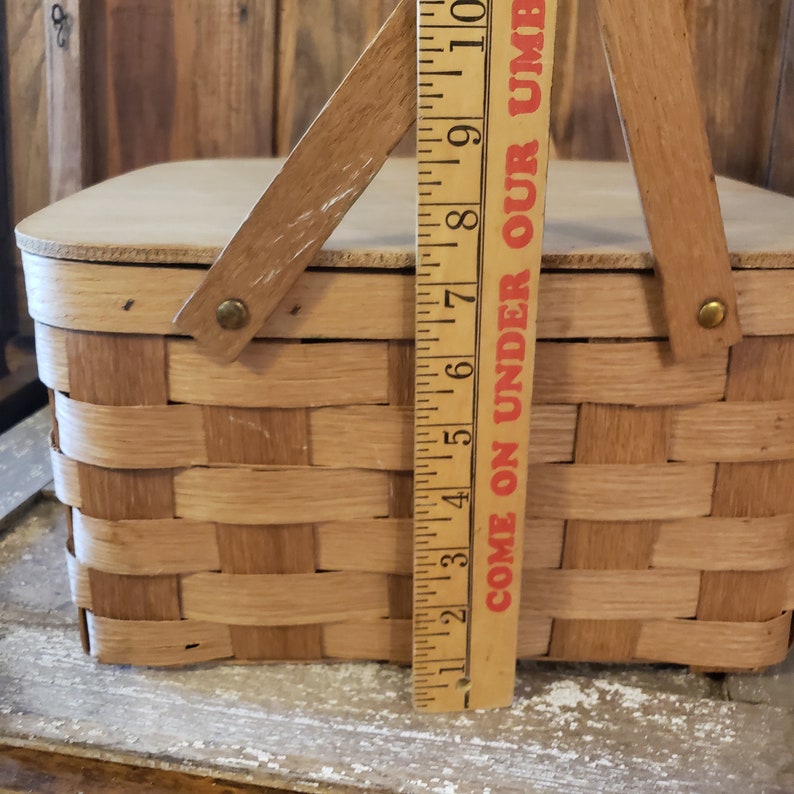 Puede incluir: Una cesta de picnic de madera con tapa abatible y dise&ntilde;o tejido. La cesta tiene dos asas y una regla a su lado que dice "Come on Under Our Umbrella".