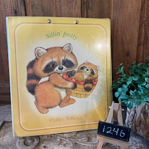 May include: A vintage children's book titled "Sittin' pretty" featuring a raccoon and baby raccoon holding a basket of red cherries. The book has a yellow cover with a brown border and the words "Critter Sitters" at the bottom.