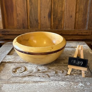 Puede incluir: Un cuenco de madera con una banda marrón oscura alrededor del borde. El cuenco está sentado sobre una superficie de madera con un diseño tallado. Una pequeña pizarra con el número 1539 escrito en ella está en el fondo.