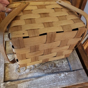 Puede incluir: Una cesta de picnic de madera tejida con dos asas largas de cuero marr&oacute;n. La cesta est&aacute; hecha de madera natural y tiene una forma cuadrada.