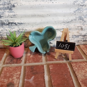 May include: A teal green ceramic shamrock-shaped dish with a speckled glaze. The dish is sitting on a brick surface next to a small wooden easel with a black chalkboard that says "1051".