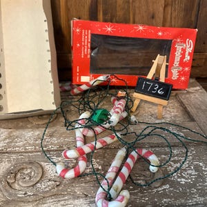 Puede incluir: Luces navideñas vintage con bombillas en forma de bastón de caramelo con rayas rojas y blancas, ensartadas en un cable verde. Se ve una caja roja con el texto "Silvestri Christmas Lights". Un pequeño caballete de pizarra muestra el número "1736".