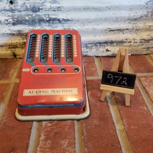 May include: A red vintage toy adding machine with blue and white accents. The machine has a total display and multiple columns of numbers. The machine is labeled "Adding Machine". A small wooden easel with a black chalkboard displaying the number "972" is in the background.