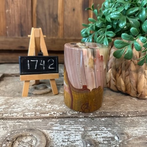 Könnte beinhalten: Ein zylindrischer, polierter Stein mit einer Mischung aus Braun-, Rosa- und Gelbtönen. Der Stein befindet sich neben einer kleinen Holzstaffelei mit der Nummer "1742" auf einer schwarzen Tafel. Eine grüne Pflanze in einem geflochtenen Korb befindet sich im Hintergrund.
