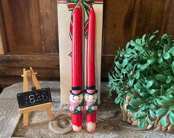 May include: Two red taper candles with snowman designs at the base, standing in front of a gift box with a red and green bow. The candles have black top hats, pink scarves, and hold green sprigs. A small wooden easel displays the number "1828".