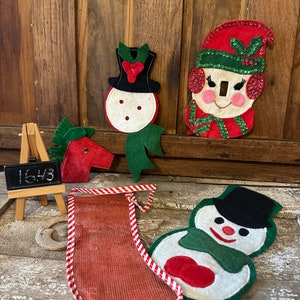 Può includere: Un set di cinque ornamenti natalizi in feltro, tra cui un pupazzo di neve, una calza, una renna e due teste di pupazzo di neve. Gli ornamenti sono decorati con feltro rosso, verde e bianco e paillettes. Gli ornamenti sono esposti su una superficie di legno.