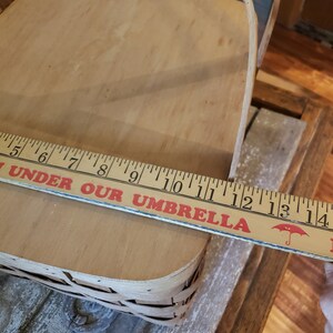 Puede incluir: Una cesta de picnic de madera con una base de mimbre tejido. La cesta mide 35,5 cm de largo y tiene una etiqueta roja y blanca que dice "Under Our Umbrella".