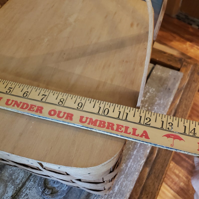 Puede incluir: Una cesta de picnic de madera con una base de mimbre tejido. La cesta mide 35,5 cm de largo y tiene una etiqueta roja y blanca que dice "Under Our Umbrella".