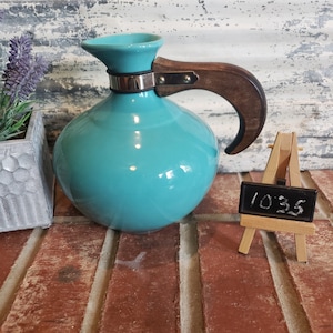 May include: A turquoise ceramic pitcher with a wooden handle and a silver band around the neck. The pitcher is sitting on a brick surface.