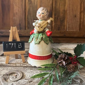 Può includere: Una campana di Natale in porcellana vintage con un bordo rosso e bianco. Una piccola statuina di angelo è seduta sulla cima della campana, suonando uno strumento musicale. La campana è circondata da verde e pigne.